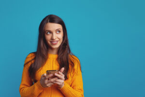 Dreamy young woman holds mobile phone has thoughtful expression thinks about received message looks away isolated over blue background. Explore the Best SMS Plugins for WordPress