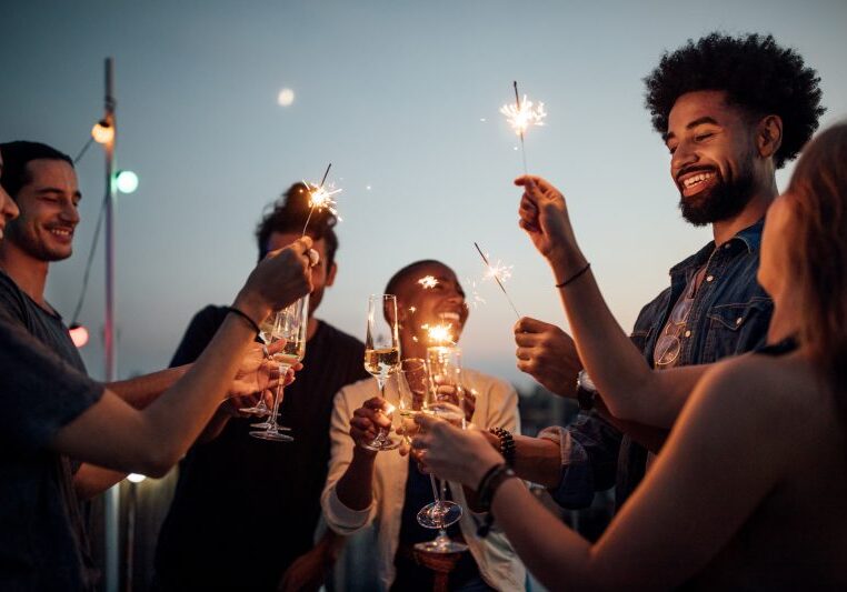 Multiracial young men and women on roof raising wineglasses and sparklers. Friends celebrating at reunion party on rooftop.