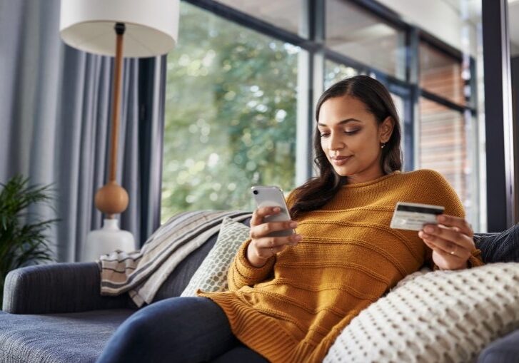 Shot of a beautiful young woman using her cellphone and credit card while relaxing on a couch at home