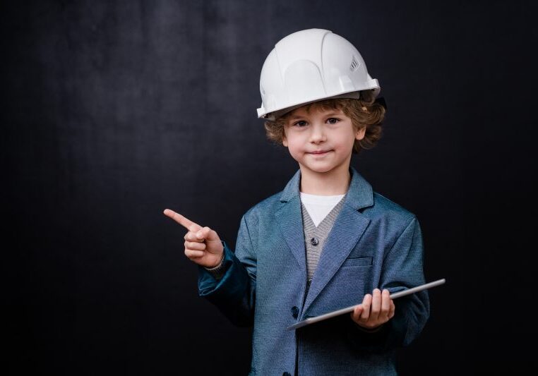 Cute little boy in hardhat and formalwear holding digital tablet while looking at you and pointing aside against black background