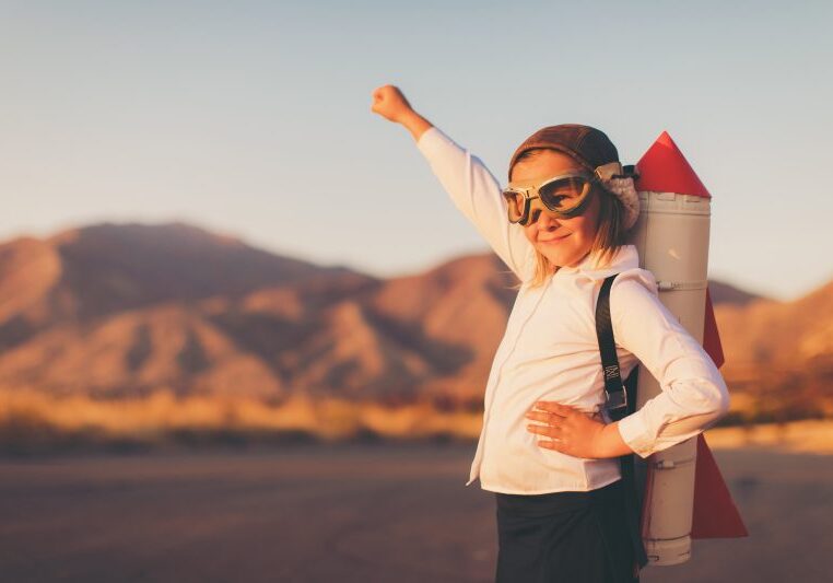A young girl businesswoman or student dressed in business attire looks to the distance while wearing a rocket strapped to her back and a flying cap and goggles. She is standing on a country road among the mountains of Utah, USA. She is confident in her abilities to lead her business and obtain her goals.